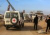 Red Cross vehicles drive through a dusty street with damaged buildings