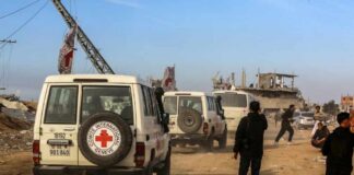 Red Cross vehicles drive through a dusty street with damaged buildings