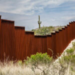 A rusty border wall in a desert landscape with a cactus in the background
