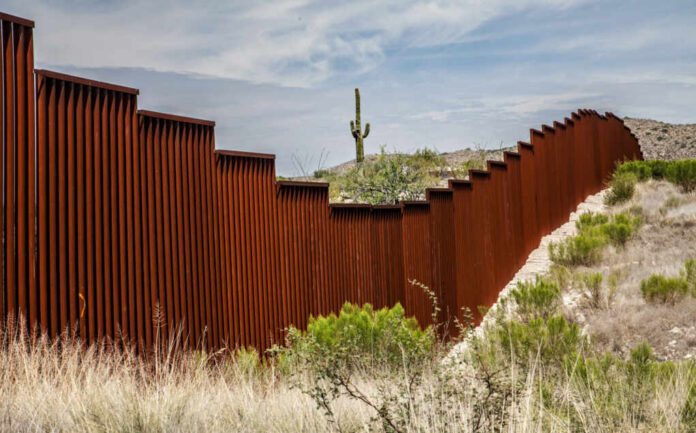 shutterstock_459602719 A rusty border wall in a desert landscape with a cactus in the background