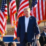 Trump Issues Warning At Rushmore A speaker at a political rally surrounded by American flags and an engaged crowd