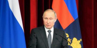 man in suit speaking at a podium with flags behind him