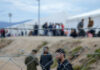 New EU Policy Stirs Refuge Debate Barbed wire fence in front of a refugee camp with people in the background