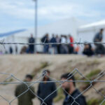 Barbed wire fence in front of a refugee camp with people in the background