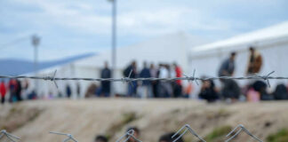 New EU Policy Stirs Refuge Debate Barbed wire fence in front of a refugee camp with people in the background