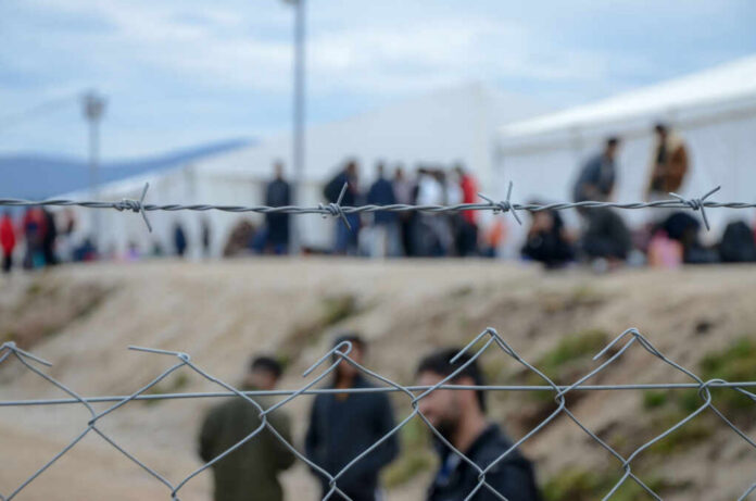 Barbed wire fence in front of a refugee camp with people in the background