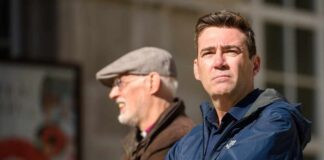 Man in blue jacket standing outdoors with another man in background