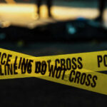 Top Politician Slain—No Suspect, No Motive Yellow police caution tape at a crime scene with blurred figures in the background