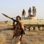 Houthis Threaten Global Oil Lifeline Soldier with RPG launcher walking past armored vehicle with troops