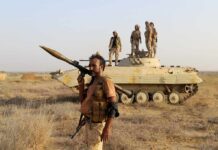 Houthis Threaten Global Oil Lifeline Soldier with RPG launcher walking past armored vehicle with troops