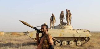 Houthis Threaten Global Oil Lifeline Soldier with RPG launcher walking past armored vehicle with troops