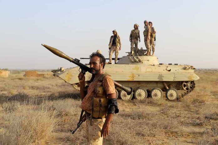 shutterstock_2391692707 Soldier with RPG launcher walking past armored vehicle with troops