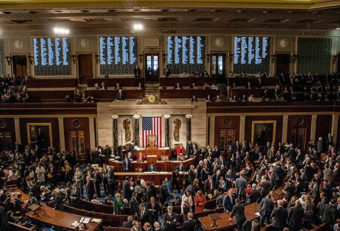 shutterstock_1186368280 Legislators gather on a chamber floor beneath a large flag