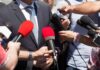 Person in suit surrounded by microphones during media interview