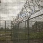 Power Clash Erupts Over ICE Site Barbed wire on chain-link fence at a prison yard