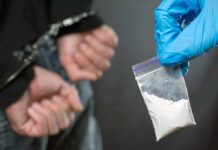 Gloved hand holds small plastic bag of white powder near handcuffed person