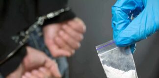 Gloved hand holds small plastic bag of white powder near handcuffed person