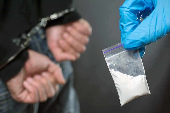 Gloved hand holds small plastic bag of white powder near handcuffed person