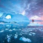 Trump Locks Greenland – Rivals Frozen Out Iceberg with arch and red-sailed boat at colorful sunset