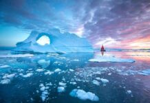 Iceberg with arch and red-sailed boat at colorful sunset