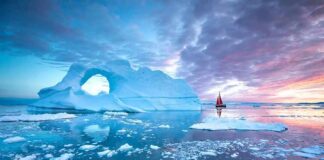 Trump Locks Greenland – Rivals Frozen Out Iceberg with arch and red-sailed boat at colorful sunset