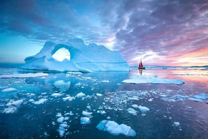 shutterstock_1209754822 Iceberg with arch and red-sailed boat at colorful sunset