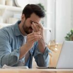 Man rubbing eyes while holding glasses at a laptop