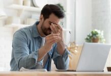 Silent Tumor Clues Revealed In Eyes Man rubbing eyes while holding glasses at a laptop