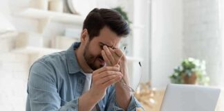 Silent Tumor Clues Revealed In Eyes Man rubbing eyes while holding glasses at a laptop