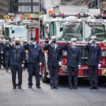 Firefighters in dress uniforms salute in front of fire trucks