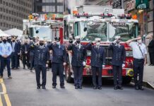 Firefighters in dress uniforms salute in front of fire trucks