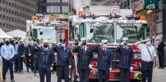 Firefighters in dress uniforms salute in front of fire trucks
