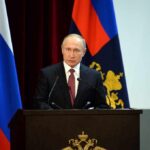 Seven Insiders Emerge In Putin Succession speaker at podium with flags in background