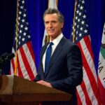 Union Fury Erupts Over Hollywood Deal Man in suit speaking at a podium with American and California flags