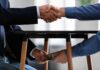 Two people shake hands while exchanging cash under a table
